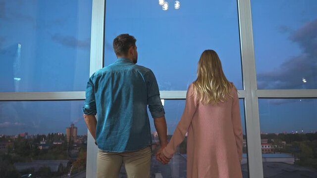 Back view of romantic couple holding hands near panoramic windows at home