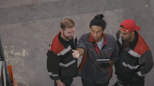 Top-view footage of serious female african american supervisor with digital tablet explaining workplan to warehouse workmen pointing at pallet racks with hand