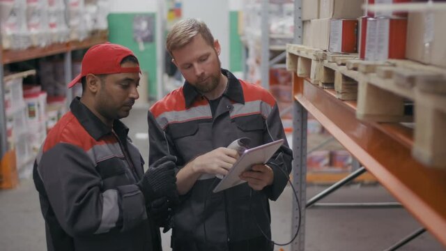 Medium shot of two multi-ethnic warehouse workmen in coveralls uniform clothing scanning barcodes on boxes on pallet racks getting data on digital tablet