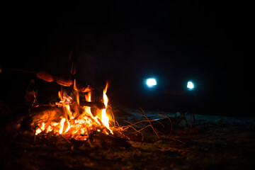 Cooking sausages on an open fire at night, in the background a car with its headlights on. Cooking...