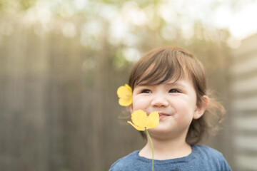 A toddler with a yellow flower on her hair holds another yellow flower in front of her face and smiles as she smells it in the garden of a house in Edinburgh, Scotland, United Kingdom