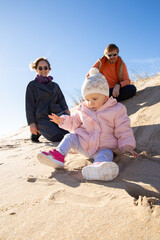 Positive mom and dad watching cute baby daughter in warm clothes, playing and rolling on sand. Leisure time and childhood concept