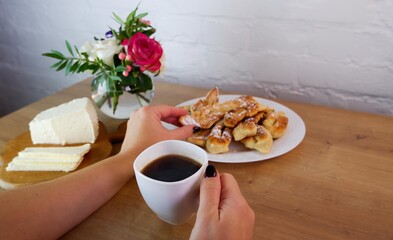 Cup of coffee in female hands, close-up. Homemade baked goods during quarantine. Homemade feta cheese on a wooden tabletop. Concept on the background of a white brick wall and a bouquet of roses.