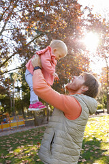 Happy dad playing with cute baby daughter in autumn park, holding and lifting kid up in air. Vertical shot. Family and childhood concept