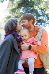 Happy parents kissing while holding cute baby girl in arms, outdoors. Medium shot, vertical. Love and family concept