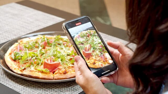 Close-up of female hands holding smartphone and making photo or video of delicious pizza in a restaurant. Blogging, technology and social media concept.