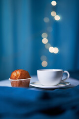 White cup of coffee with fresh muffin on the blue background with blurred lights in the cafe in evening