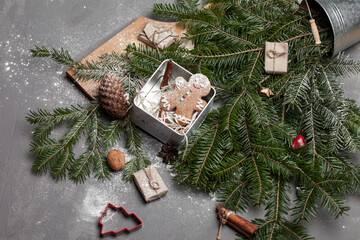 Rustic christmas composition with a metal vase with a spruce branch placed on a gray wooden board on the kitchen table at home, a box with gingerbread cookies, pine cones
