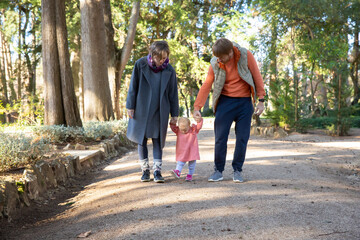 Family couple and cute baby daughter walking in autumn park. Kid holding parents hands. Front view, full length. Family and childhood concept © Mangostar