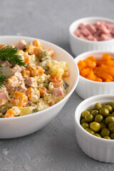 Traditional Russian Olivier salad in a white bowl and main ingredients green peas, boiled carrots, ham, pickles on a gray background. Closeup, no people