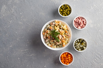 Traditional Russian Olivier salad in a white bowl and main ingredients green peas, boiled carrots, ham, pickles on a gray background. Top view, copy-space, no people