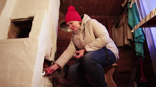 A girl in a red cap sets fire to wood in the firebox with a match. Heats the stove, prepares for Christmas. We warm the dacha in the old way. New year holiday is coming soon. Warm soft light. 4K.