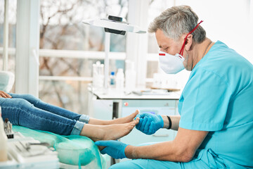Professional doctor podiatrist in medical mask examining feet and nail while patient sitting in...