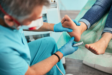 Handsome man in blue gloves holding left foot in hand while doing hard skin peeling in medical center