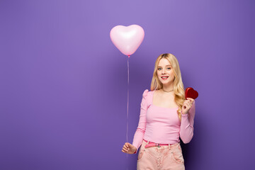 Obraz na płótnie Canvas smiling blonde young woman holding heart shaped balloon and gift on purple background.