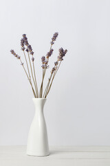 Dried lavender flowers in white vase on wooden surface over light background. Interior floral composition.