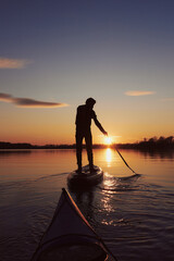 Rear view on silhouette of stand up paddle boarder paddling at sunset on river at cold time