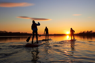 Silhouettes of the people on stand up paddle board at dusk on a flat quiet winter river with beautiful sunset colors