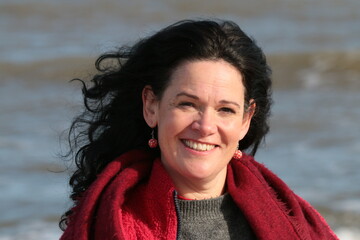 Close up portrait of attractive woman  with dark eyes black hair fair skin by sea shore on sandy beach in Winter wearing in red jacket and scarf a coast breeze, calm waves with Winter sunlight on face