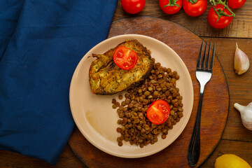 A plate of fish with lentils and tomatoes on a round stand with a fork on a blue napkin