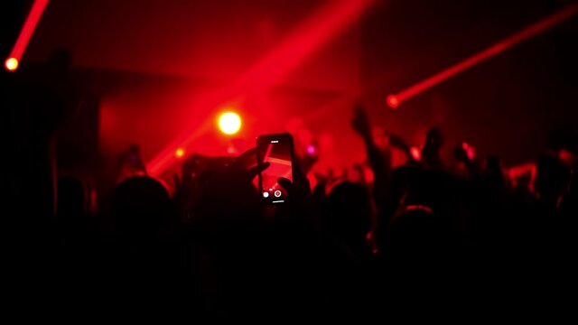 Music group gives a concert in a nightclub and fans dance in front of the stage in slowmo. People jump with hands up. Some of them are making video on mobiles. Silhouettes illuminated by flashlights.