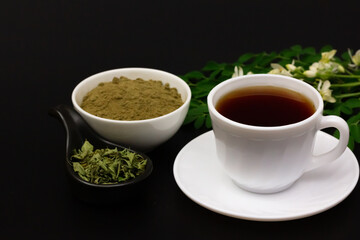 moringa tea in white cup and moringa powder in white bowl with green leaf and flower isolated on black background