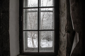 View through an old window in an abandoned house. Snowy trees outside the window. Shabby walls of an abandoned house