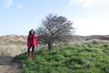 Female figure wearing mask during Covi-19 outbreak lockdown outdoors for beach walk in Autumn 2020 sunny landscape wearing red jacket, boots standing on grass by a bare tree with sand dunes background