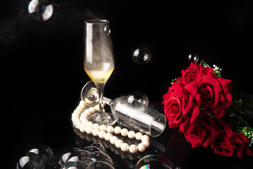 Glass of champagne, red roses, pearl necklace and soap bubbles, black background, selective focus.