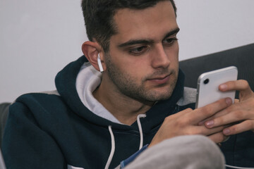 young man with mobile phone or smartphone and wireless headphones on the sofa at home