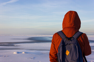 Back view of male tourist with rucksack standing on coast of the frozen bay while journey. Man traveler wearing red jacket with backpack explore nature. Wanderlust lifestyle.
