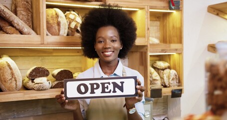 Close up portrait of cheerful young African American pretty female seller in apron holding Open sign in hands standing in small bakehouse in good mood on reopening, looking at camera and smiling