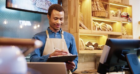 Handsome young African American male seller working in bakery shop writing in planner. Portrait of happy man taking notes in documents at workplace in own bakehouse store. Worker concept