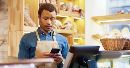 Close up of African American young handsome male worker in apron stands in bakery shop, texting and typing on smartphone. Man seller browsing online on cellphone. Business concept. Retail industry
