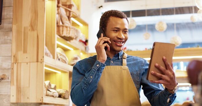 Close up of joyful African American young handsome man worker in apron stands in bakery shop, speking on smartphone and using tablet device. Business concept. Retail industry