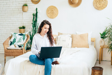 Cute young brunette business woman is sitting on the bed in the room with a laptop. Freelance work and study.