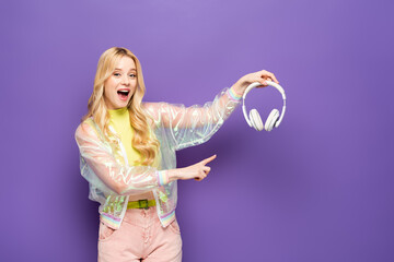 excited blonde young woman in colorful outfit pointing at headphones on purple background.