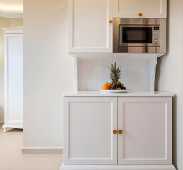 Front view of mini kitchen cupboard module cabinet with microwave and fruits plate. Classic style interior of white room with empty copy space wall