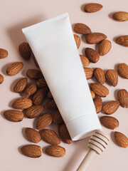 A white tube of hand cream lies on a beige background. Sprinkled with almonds and a honey stick. Cosmetics with natural ingredients. Honey and nuts in cosmetics.