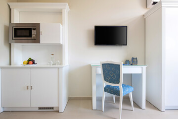 Front view of white wall with TV, table, classic blue chair, wooden kitchen cupboard cabinet with built-in appliances in small room modern interior