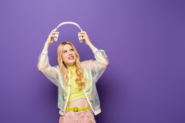 confused blonde young woman in colorful outfit and headphones on purple background.