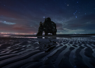 Picturesque view of starry sky with Northern lights over seashore with huge rock at night in Iceland in long exposure