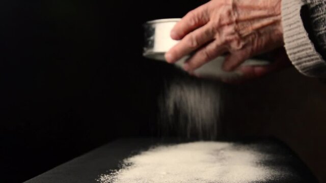 Old hands sift flour on a dark background close-up. An old mother sifting flour through a sieve on a dark background. A grandmother with a sieve in her hands sifts flour for baking bread.Bread concept