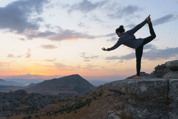 Low angle side view of flexible female standing in Lord of the Dance position against cloudy sunset sky while practicing yoga on top of rocky mountain