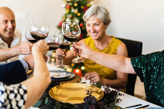 Happy mature friends gathering at festive table near shining Christmas tree and clinking wineglasses while celebrating Christmas eve together