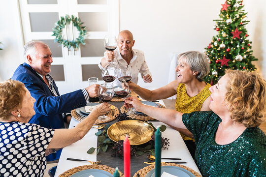Happy mature friends gathering at festive table near shining Christmas tree and clinking wineglasses while celebrating Christmas eve together