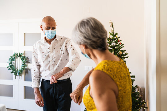 Positive Mature Couple In Smart Casual Outfits And Face Masks Greeting By Bumping Elbows While Standing Near Decorated Shiny Christmas Tree