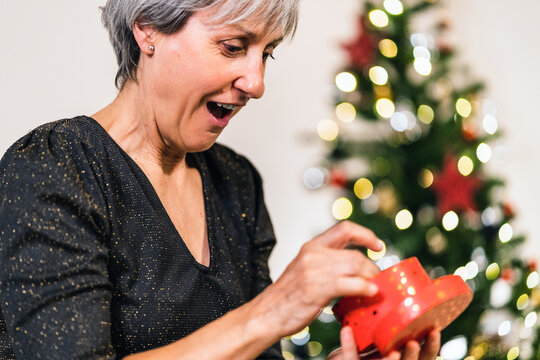 Side view of excited mature female in elegant dress opening box and enjoying gift during Christmas celebration at home