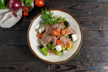 Meat salad with roast beef and feta cheese. Cold appetizer of lettuce leaves, cherry tomatoes, beef, veal or pork, feta cheese and olives in a ceramic plate on a wooden kitchen table.