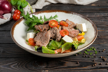 Meat salad with roast beef and feta cheese. Cold appetizer of lettuce leaves, cherry tomatoes, beef, veal or pork, feta cheese and olives in a ceramic plate on a wooden kitchen table.